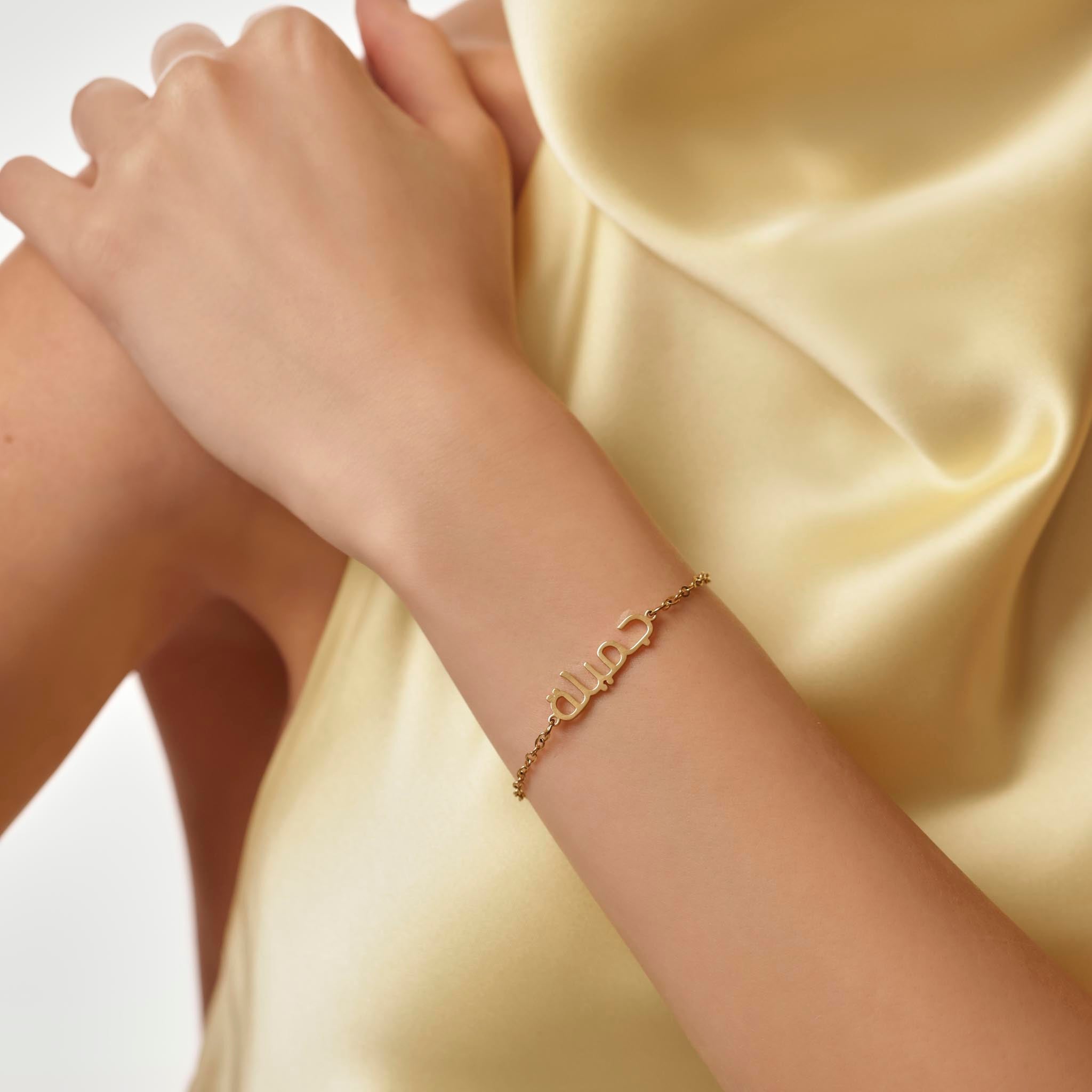 Gold arabic name bracelet on a wrist with a soft yellow fabric background