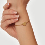 Gold bracelet with a heart-shaped tag on a wrist against a neutral background